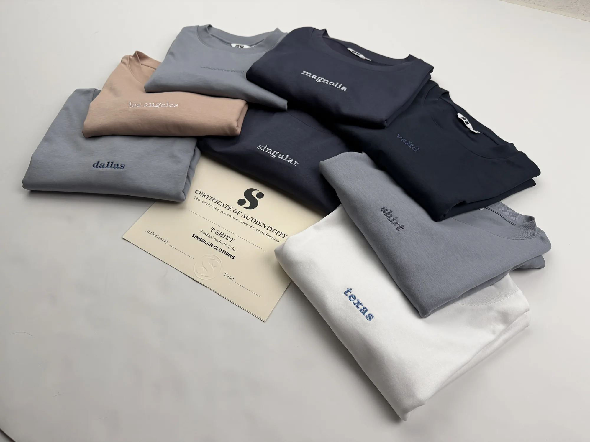 The collection seen from an angle across the studio table, the certificate of authenticity laid among folded tees