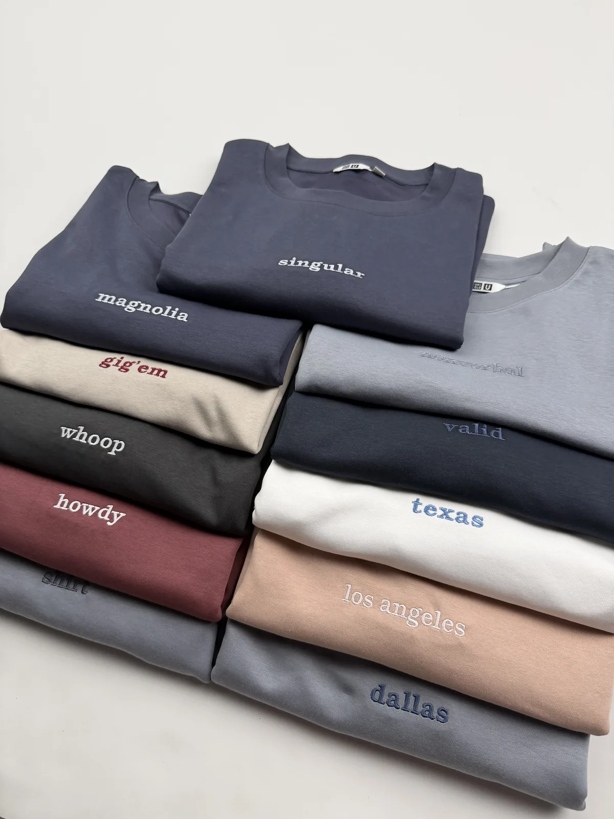 The collection stacked in two columns on the studio table — singular and magnolia in navy at the top, with gig 'em, whoop, howdy and shirt down the left and nonverbal, valid, texas, los angeles and dallas down the right