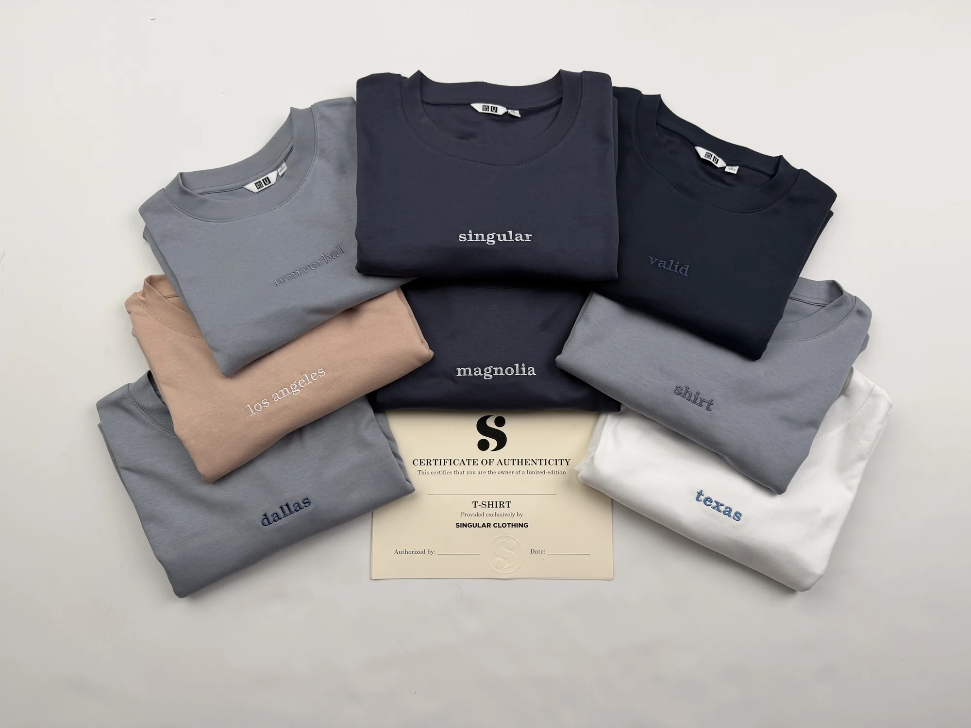 The full run of singular by syntax tees fanned out on a studio table around a certificate of authenticity — nonverbal, singular and valid across the top, los angeles, magnolia and shirt below, with dallas and texas at the edges
