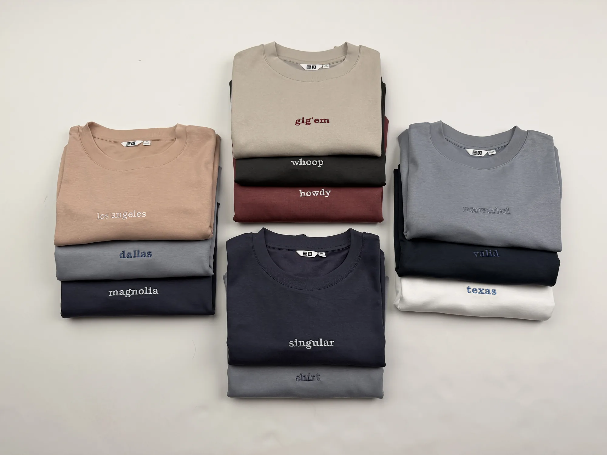 The same tees stacked in three groups on the studio table — los angeles, dallas and magnolia at the left, gig 'em, whoop and howdy in the centre, nonverbal, valid and texas at the right, with singular and shirt below